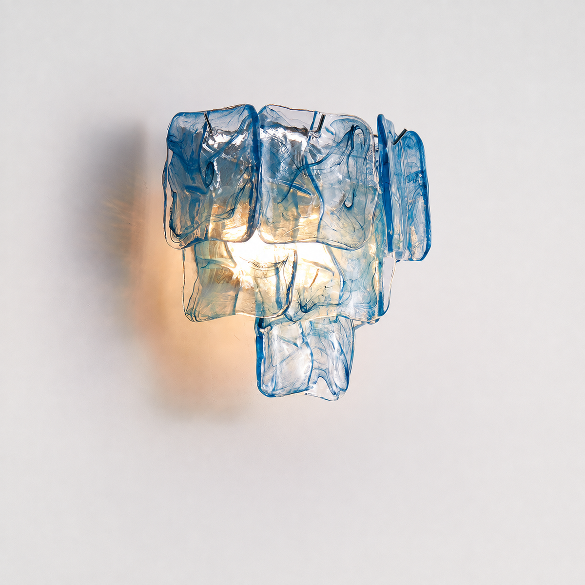 A modern wall sconce light fixture made of stacked, irregular translucent blue glass pieces, glowing with a warm light against a plain off-white background.