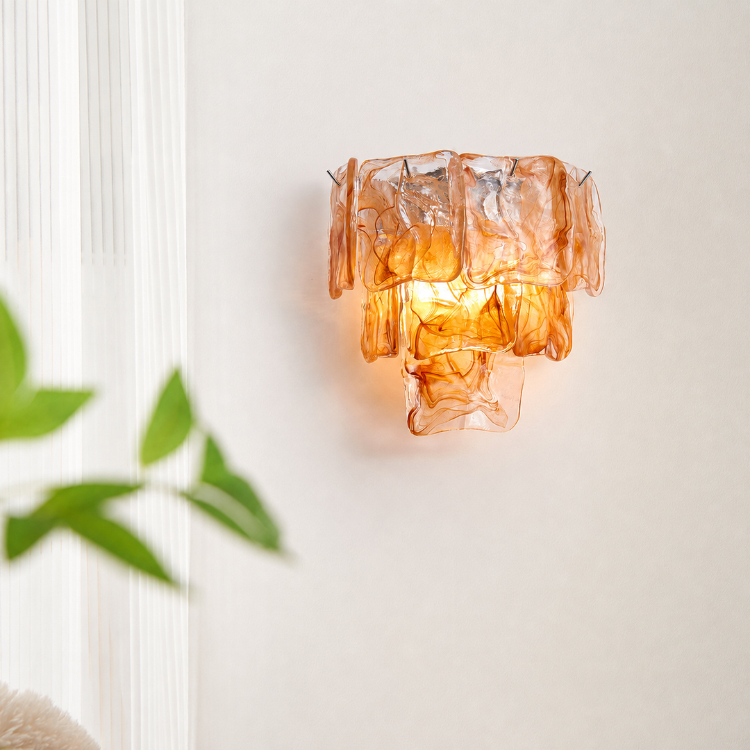 An amber-orange textured glass wall sconce mounted on a white wall, with soft green leaves blurred in the foreground on the left.