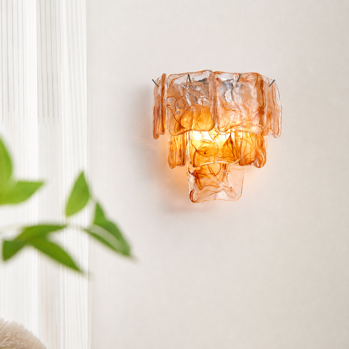 An amber-orange textured glass wall sconce mounted on a white wall, with soft green leaves blurred in the foreground on the left.