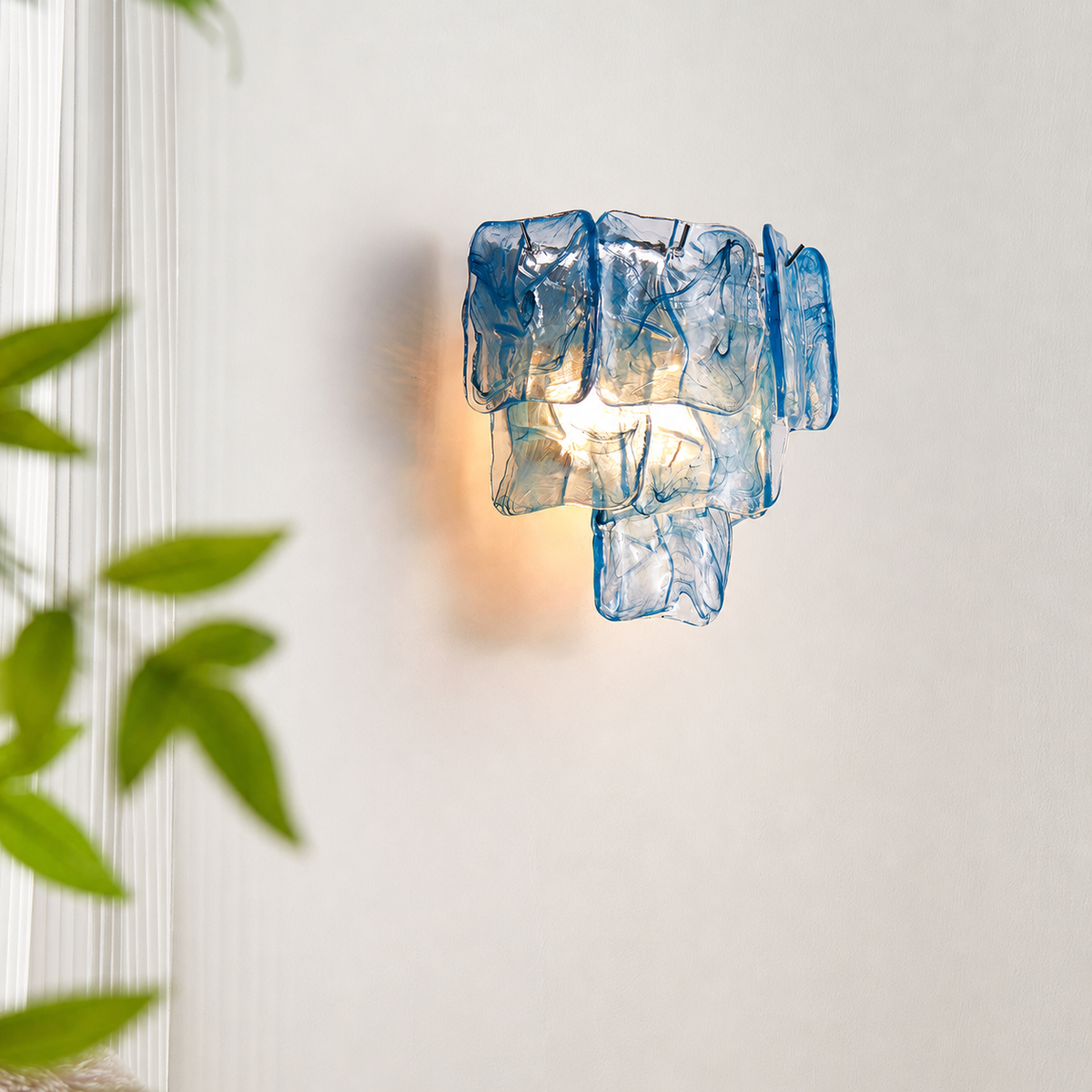 Blue glass wall sconce glowing warmly on a white wall beside decorative molding and green leaves.
