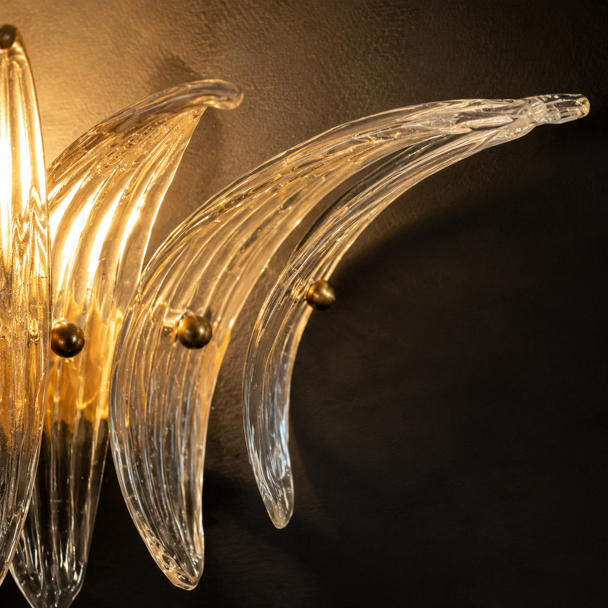 A close-up of a modern wall sconce featuring curved clear glass leaf-like blades with warm gold-toned metal accents, mounted against a dark textured wall.