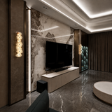 A modern living room interior featuring a large black flat-screen TV mounted against a textured beige and gray marble wall above a low cream media console. The room has dark marble flooring, warm recessed ceiling lighting, decorative wall sconces, dark curtains, and a round white bathtub visible in the foreground.