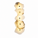 Vertical wall sconce with five overlapping cream alabaster petals, brass-tone circular accents, and a warm glow against a white background.