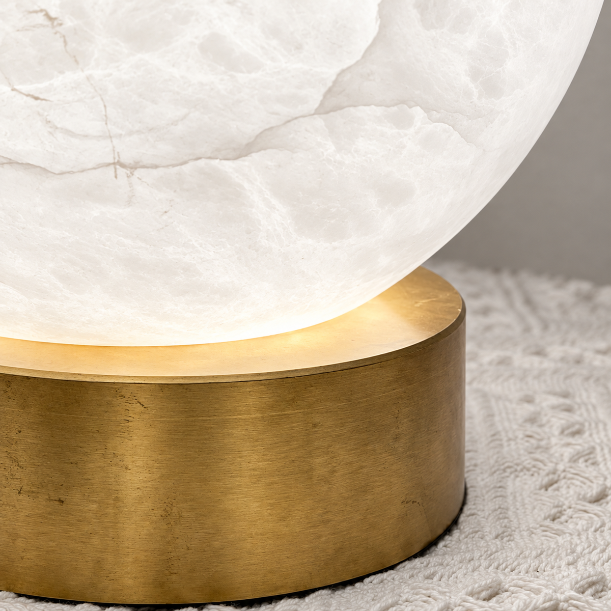Close-up of the table lamp showing its glowing white alabaster globe and brushed gold-toned copper base on textured cream fabric.