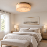 A modern flush-mount ceiling light with a textured beige fabric drum shade and a brass-toned metal rim, installed in a beautifully styled bedroom with a bed, nightstands, and lamps.