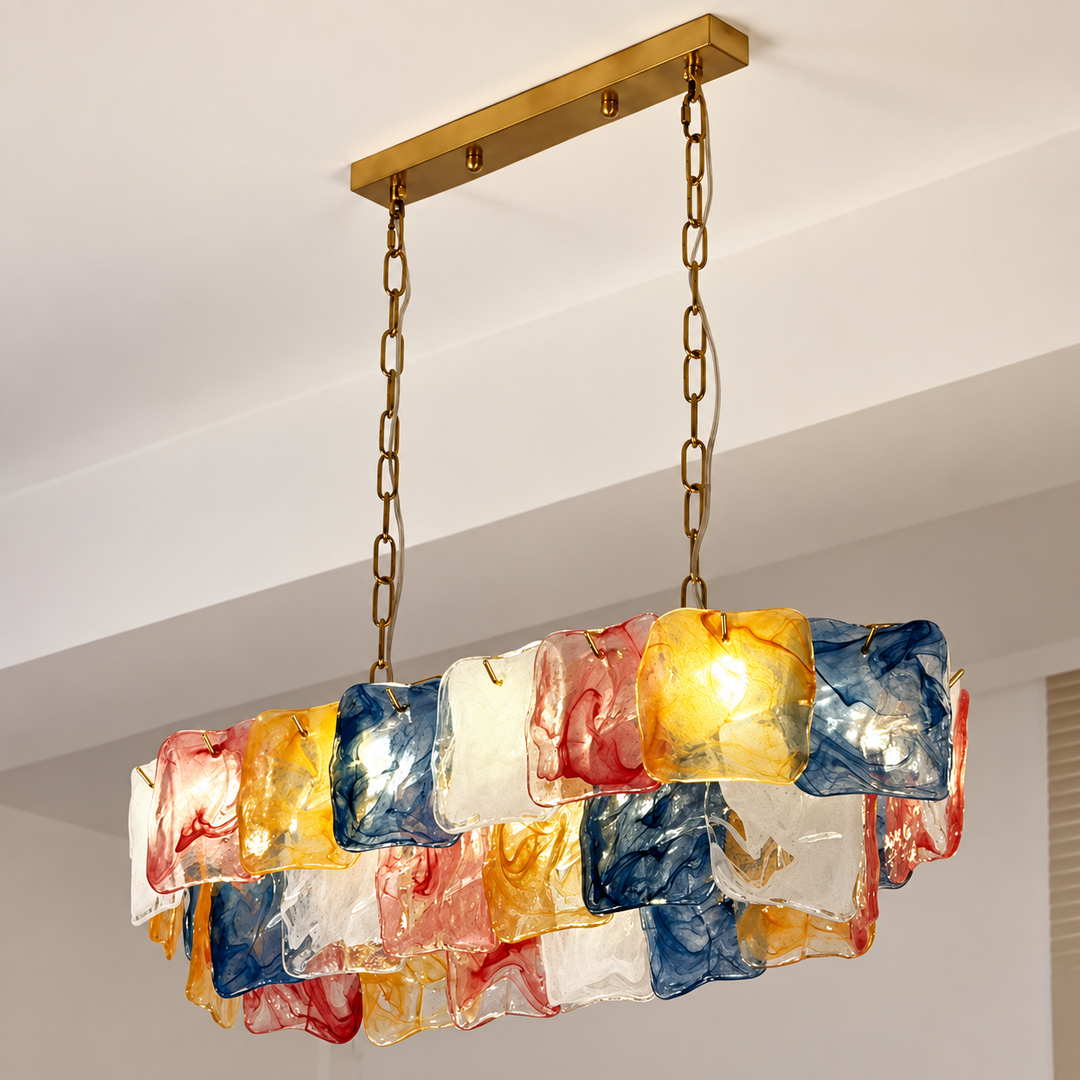 An illuminated linear chandelier features layered yellow, blue, red, and clear glass panels suspended from gold-toned chains beneath a white ceiling.