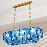 A blue glass linear chandelier hangs beside window blinds, with layered wavy panels, gold-toned chains, and a matching ceiling canopy.