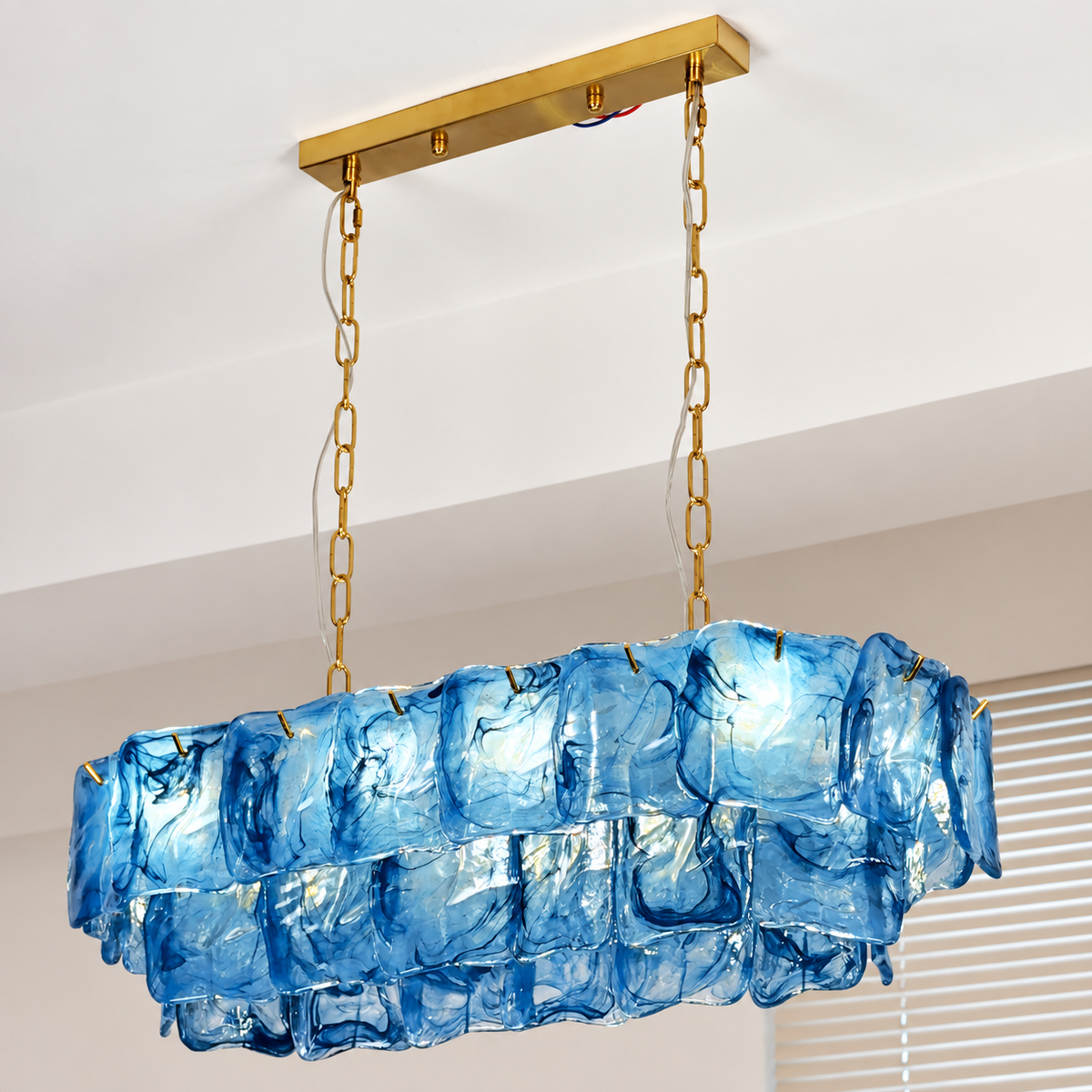 A blue glass linear chandelier hangs beside window blinds, with layered wavy panels, gold-toned chains, and a matching ceiling canopy.