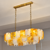 Linear chandelier with amber and frosted glass panels glows beneath a gold-toned ceiling canopy beside window blinds.
