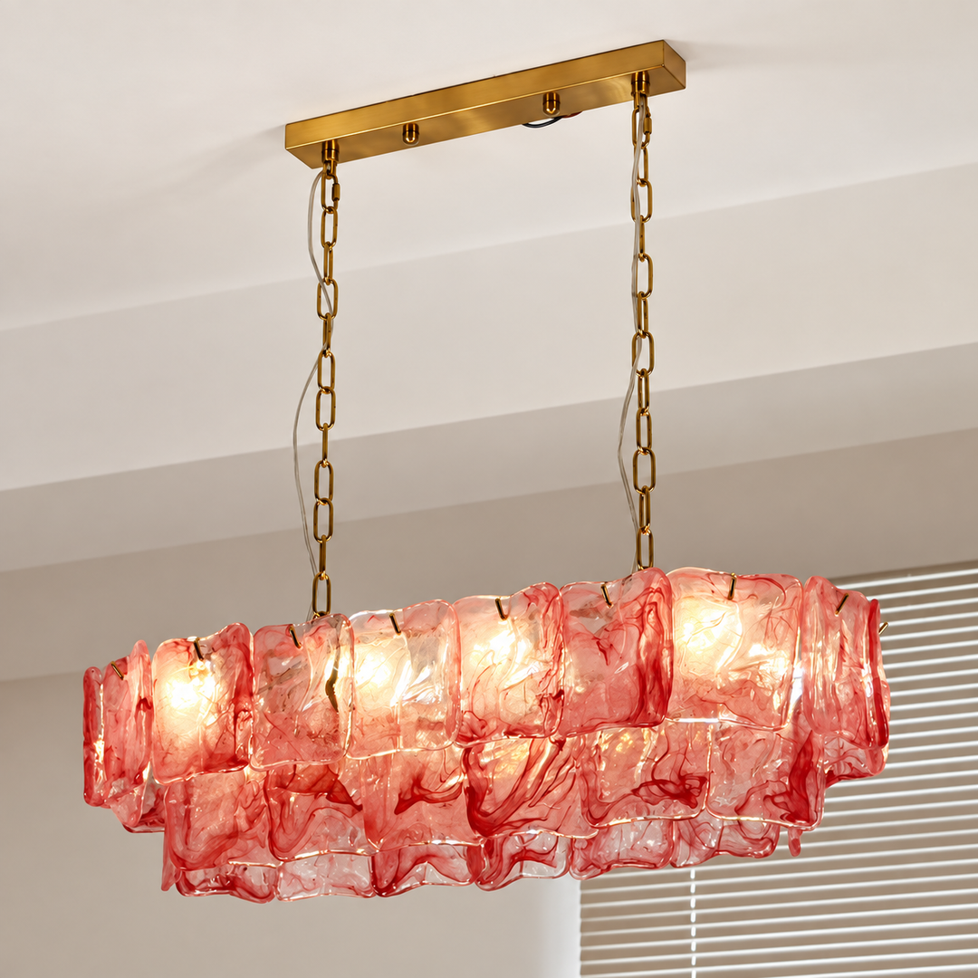 An illuminated linear chandelier with layered red textured glass panels and gold-toned chains hangs beside window blinds.