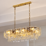 A linear chandelier glows through layered clear textured glass panels, suspended from gold-toned chains beside window blinds.