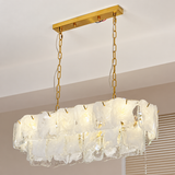 Linear chandelier with layered clear and frosted glass panels hangs from gold-toned chains beside window blinds.