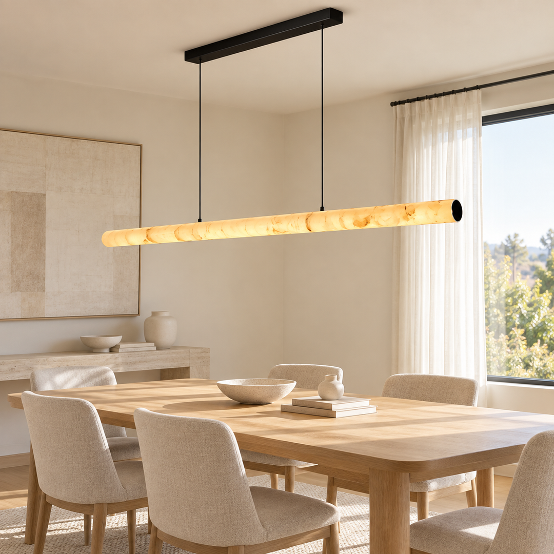 A modern linear pendant light with a warm glowing bubble or shell-like textured glass shade hangs over a light wood dining table in a bright, minimalist dining room with beige chairs and neutral decor.