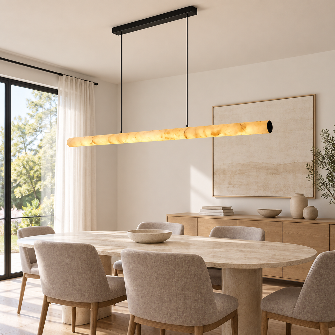 A modern linear pendant light with a warm glowing amber glass shade hangs over a dining table in a bright, contemporary dining room with beige chairs and a wood sideboard.