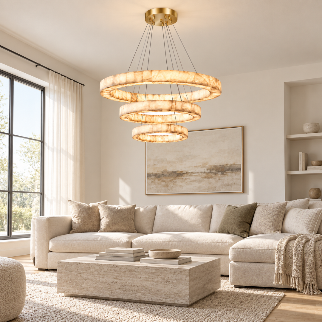 A modern multi-tiered circular chandelier with a warm gold finish and glowing textured glass rings, hanging in a bright, contemporary living room with a beige sectional sofa and light wood coffee table.