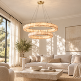 A modern two-ring double-hoop chandelier with a warm brushed gold finish and textured cream stone-like rings, hanging in a bright, modern living room with a beige sofa and marble coffee table.