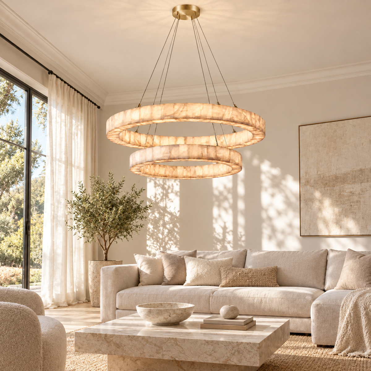 A modern two-ring double-hoop chandelier with a warm brushed gold finish and textured cream stone-like rings, hanging in a bright, modern living room with a beige sofa and marble coffee table.