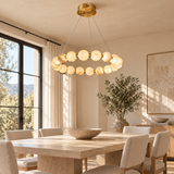 An alabaster ring chandelier with gold-tone metal hangs above a rectangular dining table in a sunlit room with cream upholstered chairs.