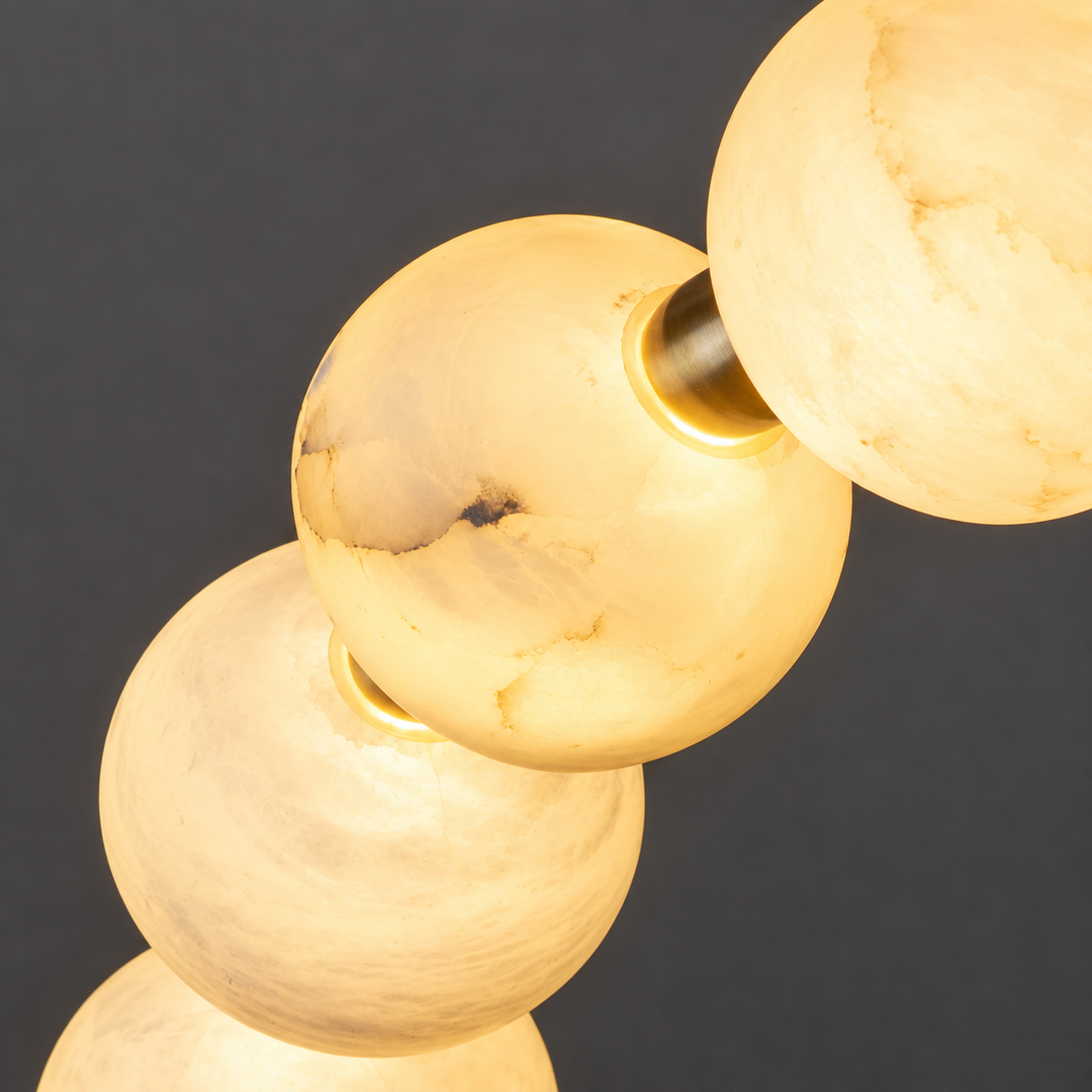 A close-up of the ring chandelier shows glowing cream alabaster with visible veining and gold-tone metal against a dark gray background.