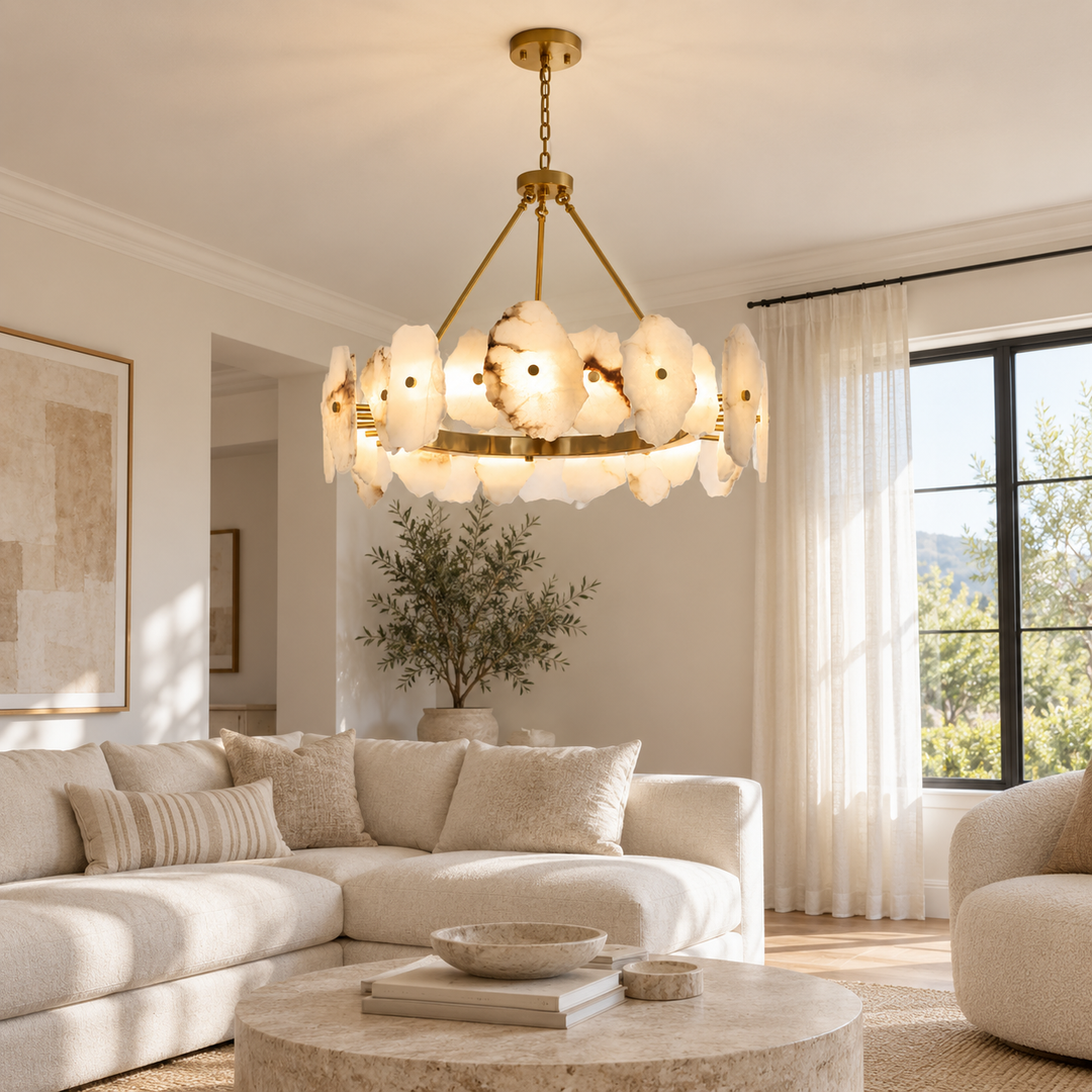 A modern chandelier with a gold brass frame and multiple organic-shaped white glass shades hangs in a bright, contemporary living room with a beige sectional sofa and large windows.