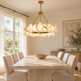 A modern chandelier with glowing white petal-shaped glass shades hangs over a light stone dining table and upholstered chairs in a bright, sunlit dining room.