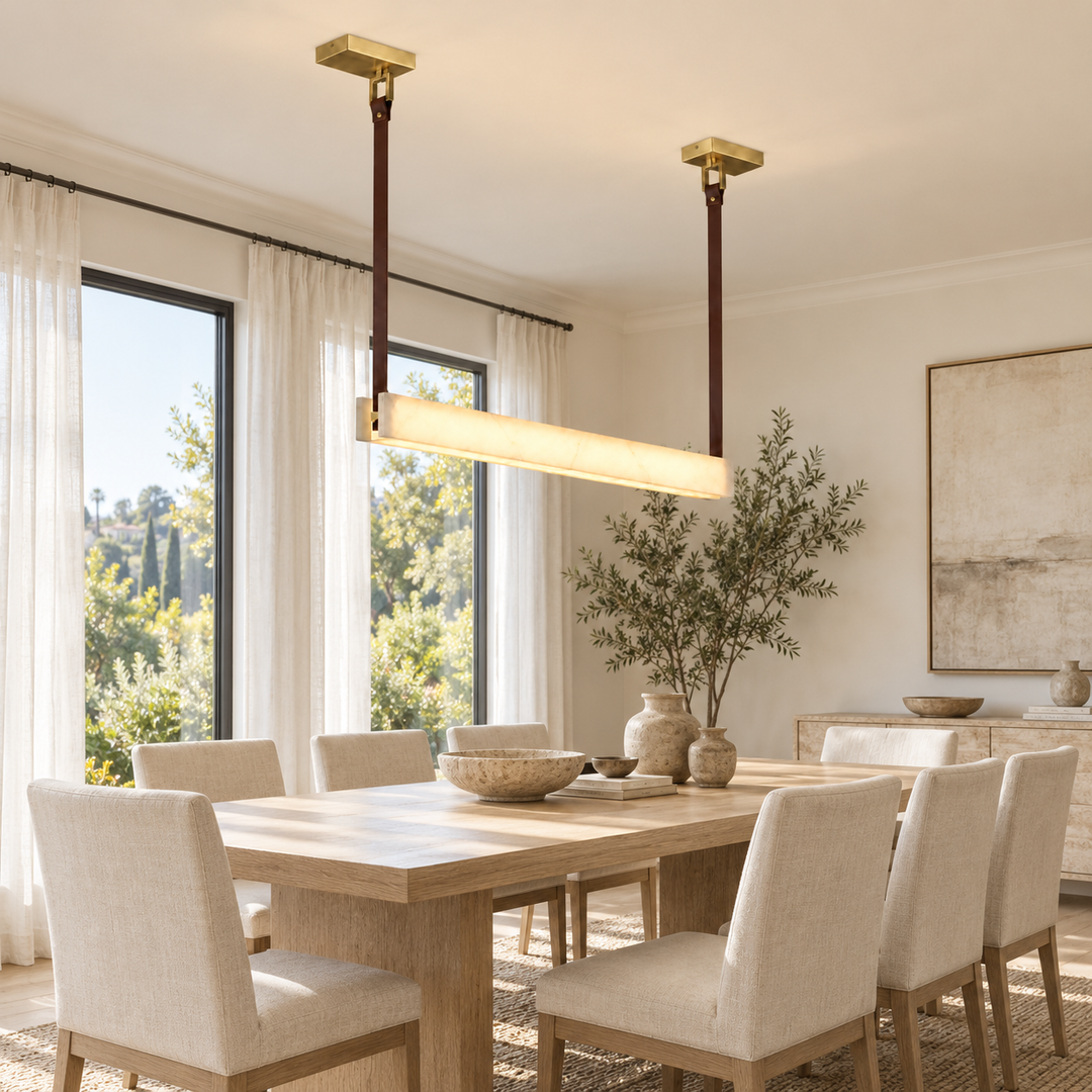 A modern rectangular linear pendant light with a warm white glowing bar and gold-toned ceiling mounts, hanging over a light wood dining table in a bright, contemporary dining room with beige upholstered chairs.