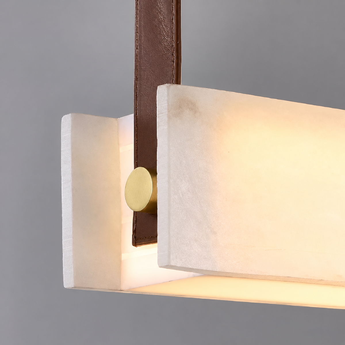 Close-up of a glowing white alabaster linear pendant light with a brown leather strap and round gold-tone fastener against a gray background.