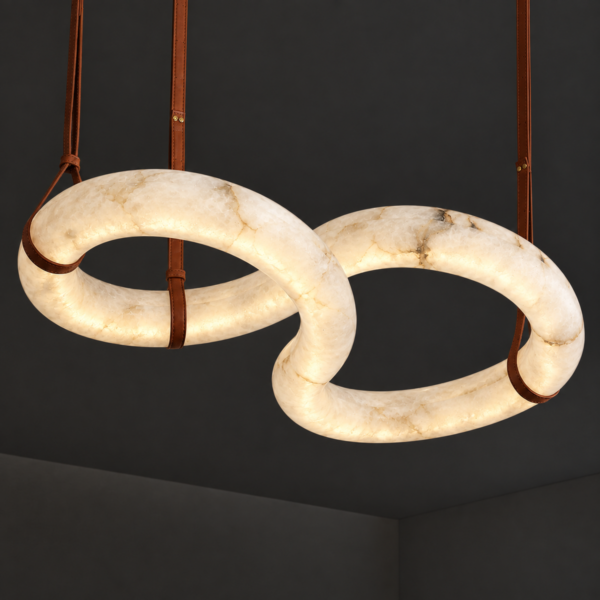 A modern two interlocking oval ring pendant chandelier made of warm white marble with brown leather straps, shown hanging against a dark gray ceiling.
