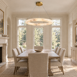 Illuminated cream alabaster ring chandelier hangs above a rectangular wooden dining table surrounded by upholstered chairs in a bright dining room.