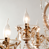 Close-up of a decorative chandelier with clear glass candle-style bulbs, gold-toned metal leaf details, and hanging crystal prisms against a light neutral background.
