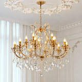 An ornate gold chandelier with multiple candle-style lights and hanging crystal prisms, suspended from a decorative ceiling in a bright, elegant room with white walls and curtains.