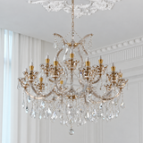 An ornate multi-light crystal chandelier with gold-toned metal arms and hanging clear glass prisms, suspended from a decorative ceiling in a bright white room.