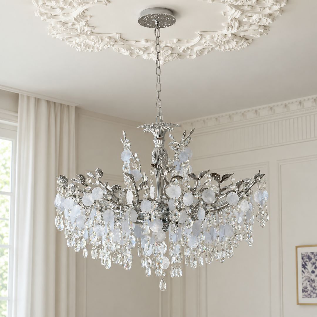 A silver-toned crystal chandelier with clear and frosted glass droplets, hanging from an ornate white ceiling medallion in a bright, classic room with cream walls and curtains.