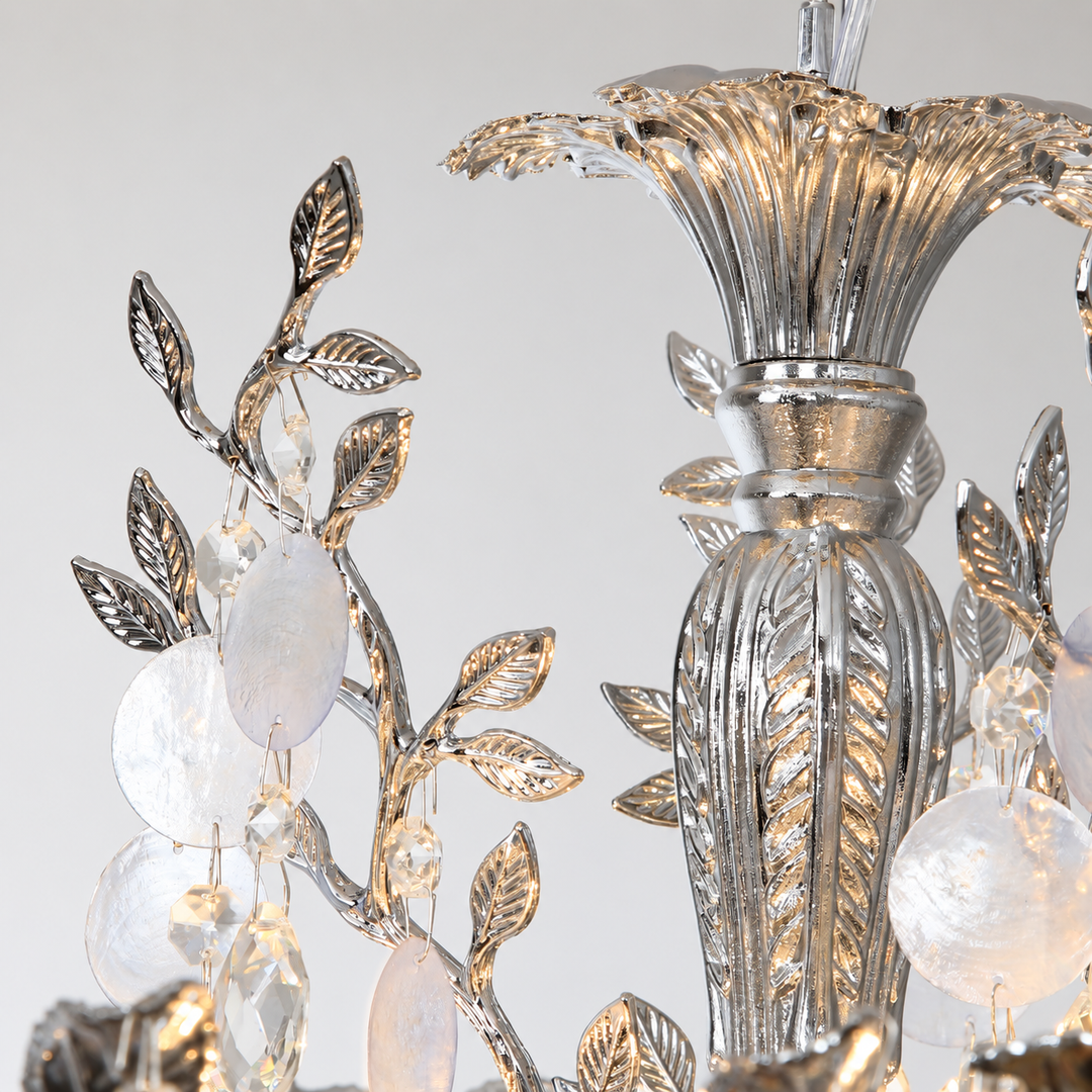 Silver-toned metal chandelier detail with leaf and vine motifs, clear faceted crystal drops, and frosted oval glass accents against a light neutral background.