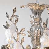Silver-toned metal chandelier detail with leaf and vine motifs, clear faceted crystal drops, and frosted oval glass accents against a light neutral background.