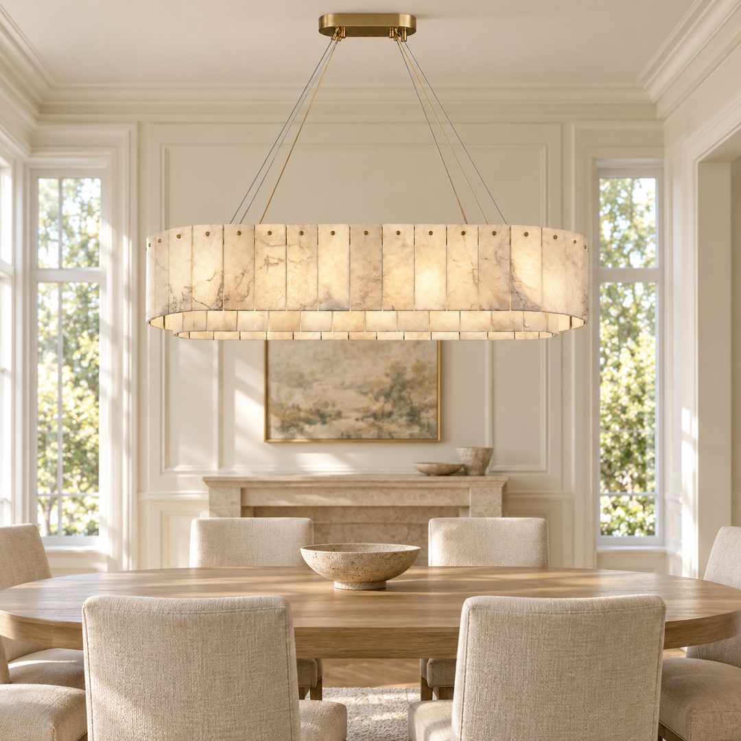 A large rectangular chandelier with a warm glowing marble-look shade hangs over a dining table in a bright, elegant dining room with cream walls, tall windows, and upholstered chairs.