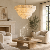 Tiered chandelier with warm glowing alabaster discs hangs above a wooden coffee table in a neutral living room with cream armchairs.