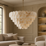 A large chandelier with overlapping oval white and cream stone-like discs hangs in a warm beige living room with a sofa, armchair, and built-in shelves.