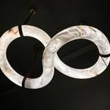 A modern infinity-shaped chandelier made of glowing white onyx with natural brown veining, suspended by dark straps against a dark ceiling.