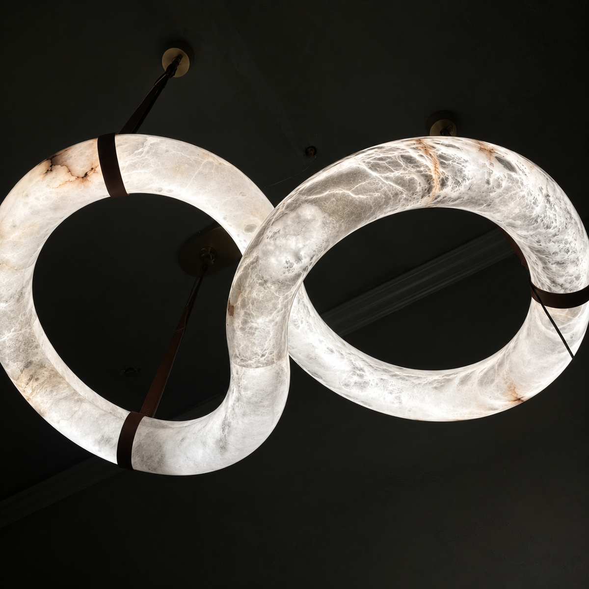 A modern infinity-shaped chandelier made of glowing white onyx with natural brown veining, suspended by dark straps against a dark ceiling.