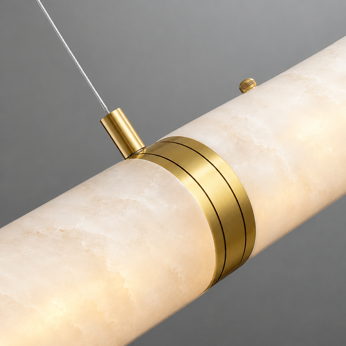 Close-up of a linear pendant with glowing cream alabaster, gold-toned detailing and a suspension cable against a grey background.