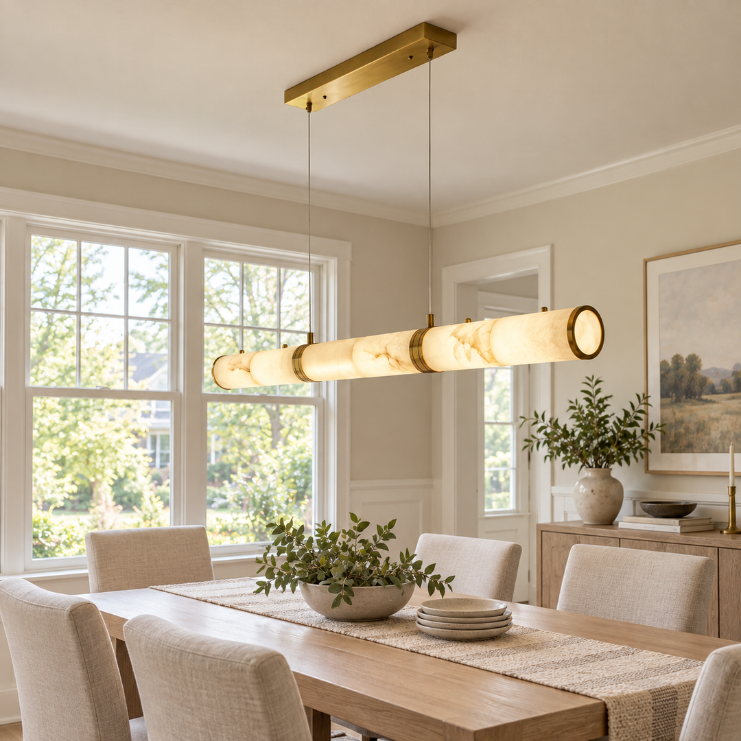 A modern linear pendant light with a warm brass frame and cylindrical cream marble-look shades hangs over a dining table in a bright, neutral-toned dining room.