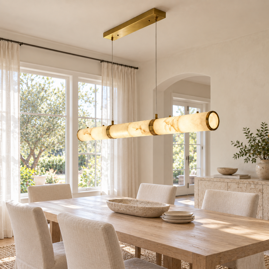 A modern linear pendant light with a brushed gold metal frame and cylindrical warm-white stone or acrylic shades, hanging over a dining table in a bright, neutral-toned dining room.