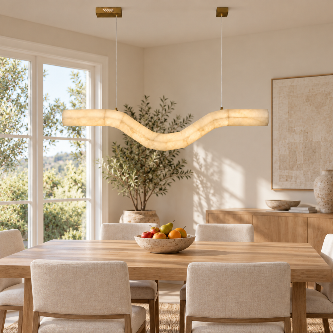 A modern wavy pendant light with a warm cream-colored textured shade hangs over a wooden dining table in a bright, neutral-toned dining room with upholstered chairs and a potted olive tree.