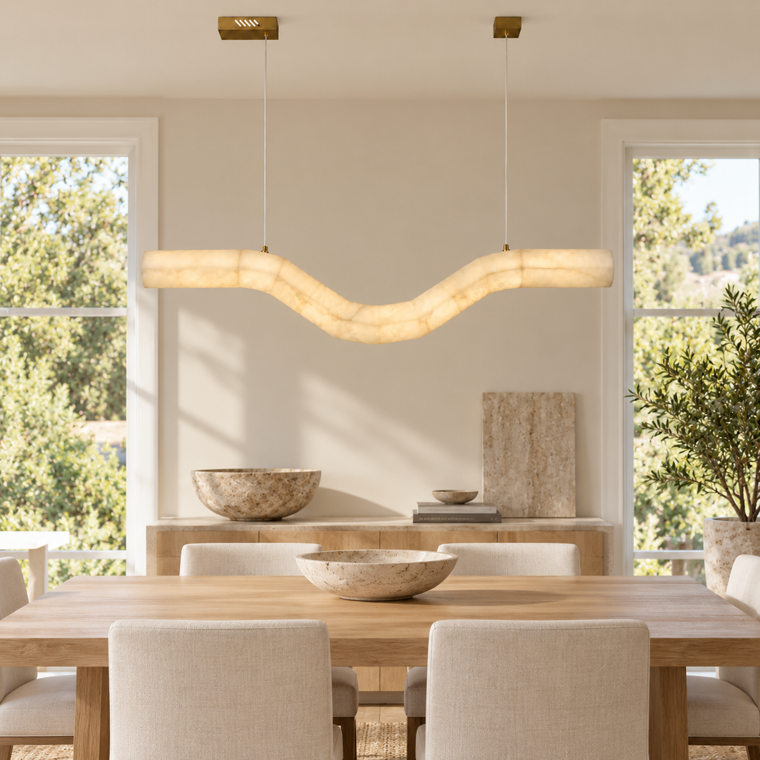A modern wavy pendant light with a warm cream-colored textured shade hangs over a wooden dining table with upholstered chairs in a bright, contemporary dining room.