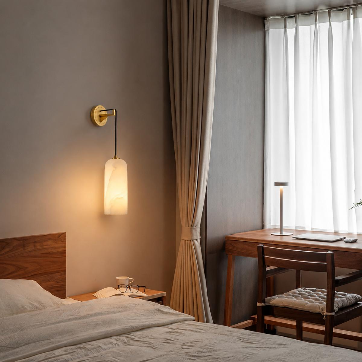 A modern wall-mounted sconce with a cylindrical frosted glass shade and a gold-toned wall mount, shown illuminated in a styled bedroom setting next to a bed and desk.
