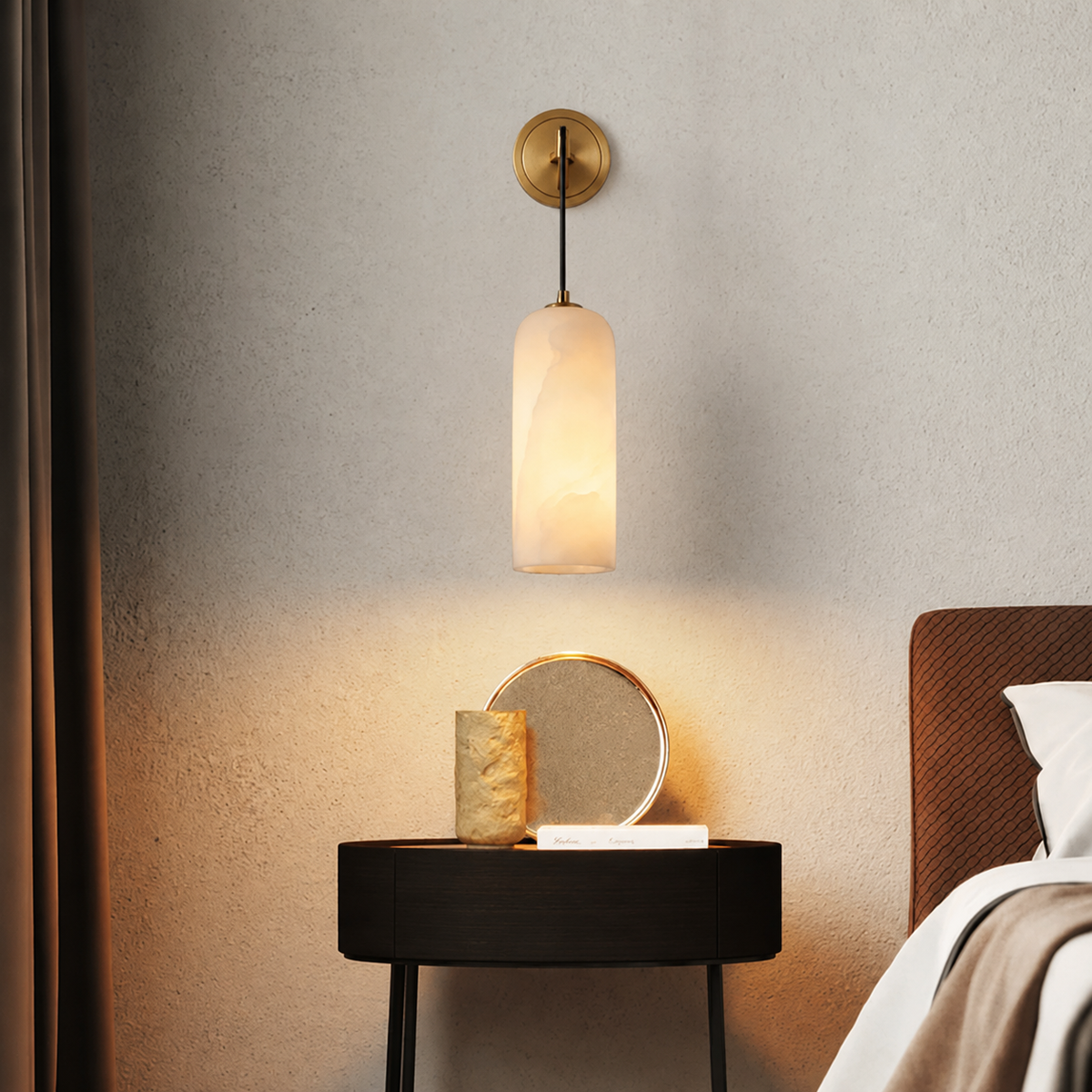 An illuminated wall sconce with a cylindrical alabaster shade and round brass mount hangs above a black bedside table beside a bed.