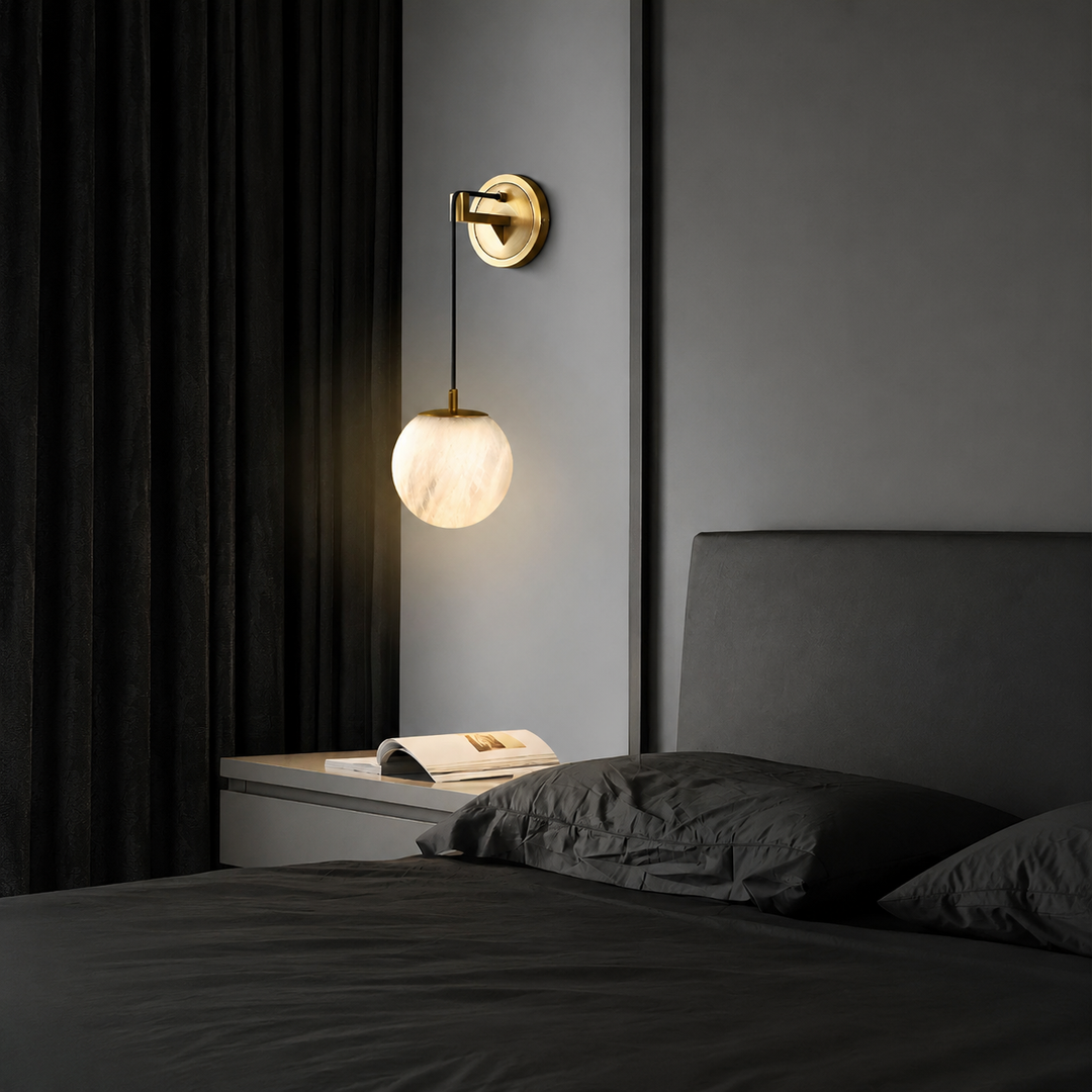 Wall pendant with a glowing alabaster globe and brass mount above a bedside table beside dark bedding and curtains.