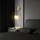 Wall pendant with a glowing alabaster globe and brass mount above a bedside table beside dark bedding and curtains.