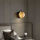 A modern wall-mounted sconce with a glowing amber glass globe and a dark metal frame, installed above a bed in a contemporary bedroom.